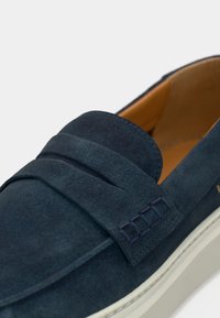 Κοντινό πλάνο ενός μοκασίνι από μπλε σουέντ με λουρί και λευκή ελαστική σόλα, που διαθέτει ταμπά εσωτερική επένδυση από δέρμα.