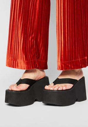 Pies con chanclas negras de plataforma combinadas con pantalones plisados de pierna ancha y color rojo brillante sobre un fondo neutro.