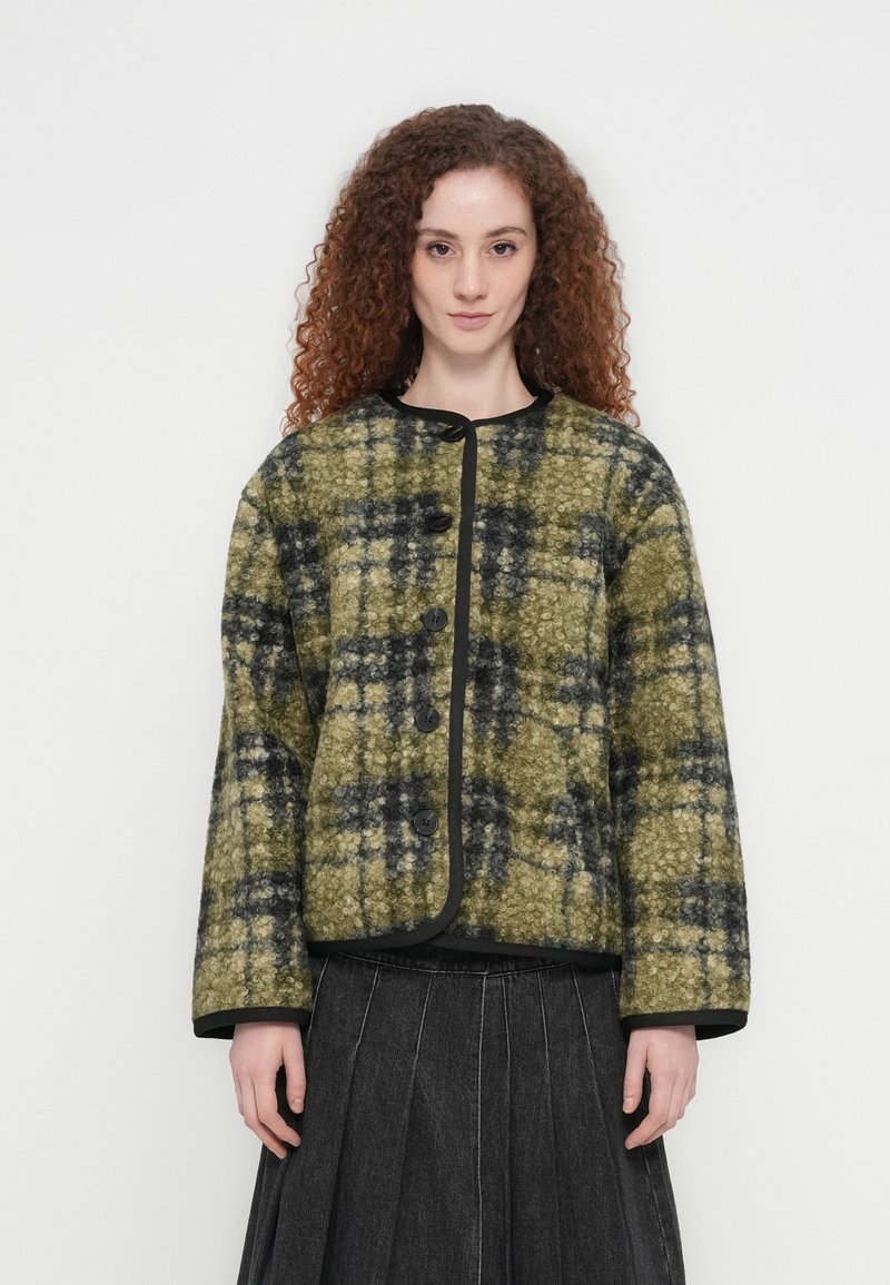 Γυναίκα με πράσινο και μαύρο υφασμάτινο καρό σακάκι με μεγάλα κουμπιά, φορεμένο πάνω από μαύρη πλισέ φούστα, που στέκεται μπροστά σε απλό φόντο.