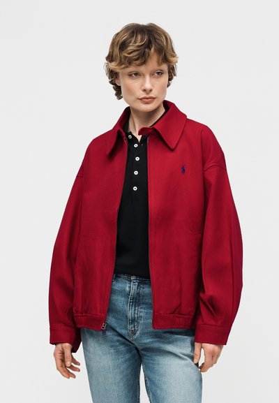 Άτομο που φοράει κόκκινο μπουφάν με φερμουάρ πάνω από ένα μαύρο πουκάμισο με κουμπιά και μπλε τζιν, που στέκεται μπροστά σε ένα απλό ανοιχτό γκρι φόντο.