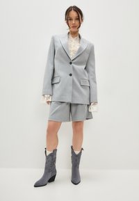 Sacou supradimensionat gri cu dungi, pantaloni scurți asortați și cizme din suede gri cu toc, vârf ascuțit și detalii decorative de cusătură.
