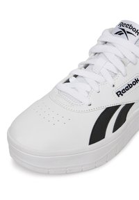 Fehér sneaker fekete kiemelésekkel, perforált orral, texturált gumitalppal és lapos fűzőkkel. Oldalán és a nyelvén a Reebok logó található.
