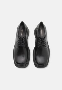 Sapatos de couro preto com uma textura suave, design de biqueira quadrada e fecho de cordões. Apresenta uma marcação discreta na sola.