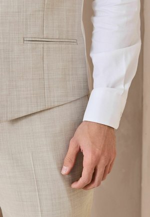 Nahaufnahme einer Hand einer Person in der Nähe der Seitentasche einer beigen Anzugjacke, die ein weißes Hemd mit geknöpftem Manschetten trägt.