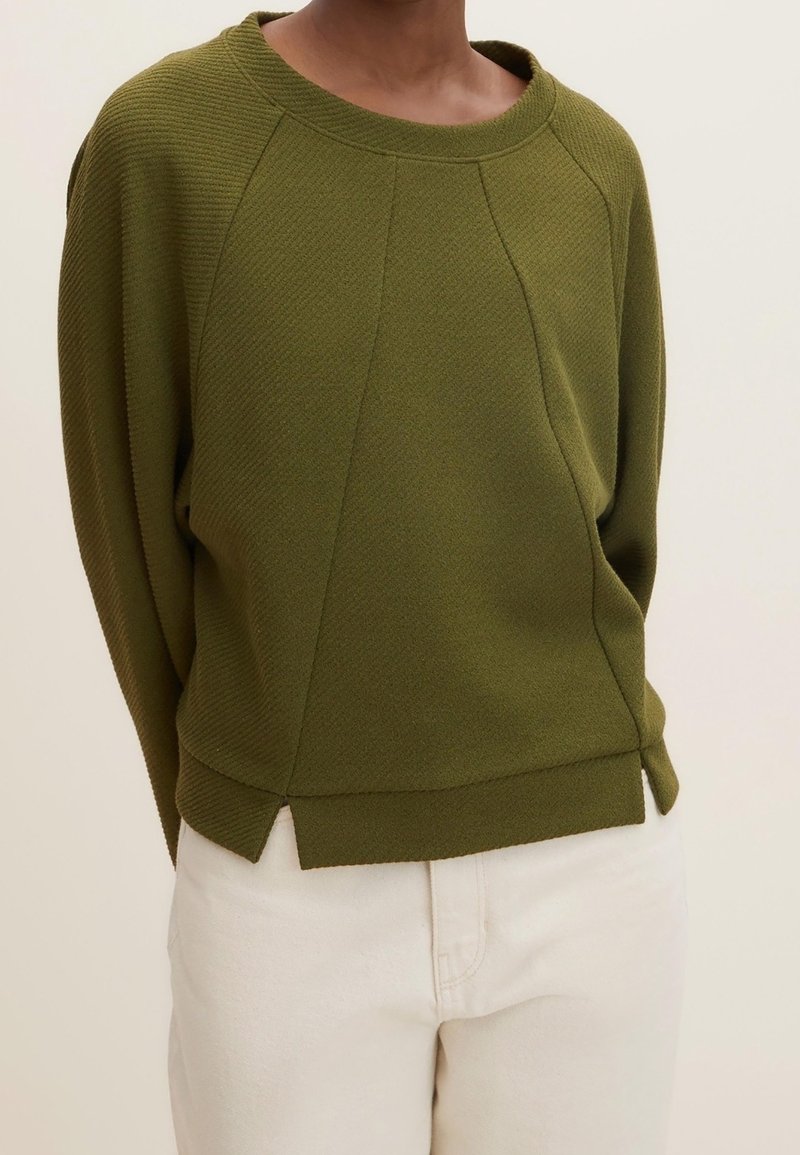 Personne portant un pull texturé vert olive à manches longues et un pantalon blanc sur un fond uni.