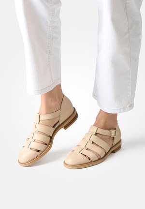 Béžové kožené sandály s uzavřenou špičkou, více pásky a přezkou, nošené s bílými zkrácenými kalhotami na bílém pozadí.