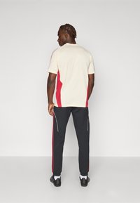 Beige Sportshirt für Herren mit roten Seiteneinsätzen, kombiniert mit schwarzen Hosen, die rote Streifen und weiße Paspeln an den Seiten aufweisen.