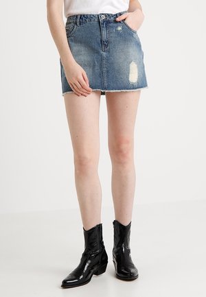 Personne portant une mini-jupe en jean bleu délavé avec un ourlet brut, un haut blanc et des bottes en cuir verni noires mi-mollet, debout sur un sol blanc.