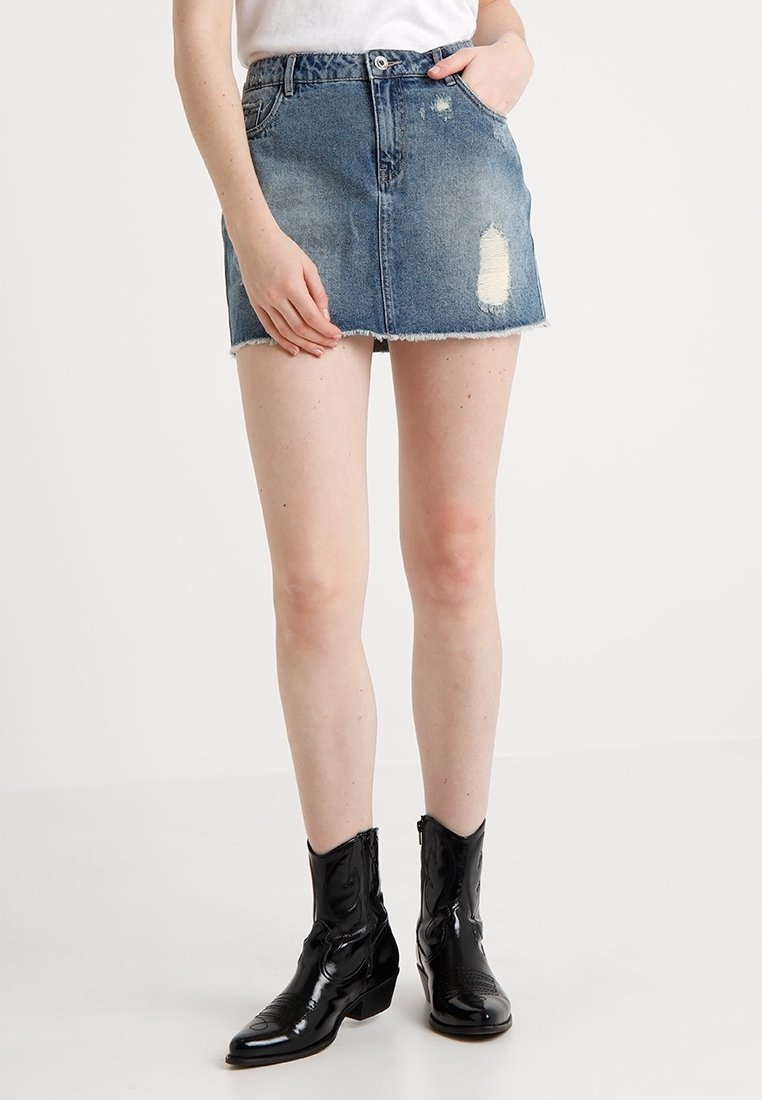 Personne portant une mini-jupe en jean bleu délavé avec un ourlet brut, un haut blanc et des bottes en cuir verni noires mi-mollet, debout sur un sol blanc.