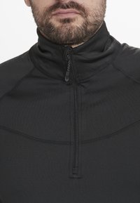 Sort zippered pullover med høj krave. Har glat stof, stikninger og en lille lynlås. Tætsiddende pasform.