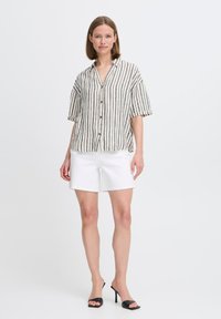 Camicetta a maniche corte a righe con bottoni, presenta una texture simile al lino in crema, marrone e verde scuro, abbinata a pantaloni bianchi e tacchi neri.
