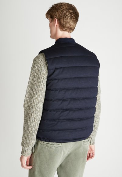 Marineblauer Pufferweste mit gesteppter Gestaltung, kombiniert mit einem hellgrauen Strickpullover. Texturierte Akzente, tailliert, klassischer Kragen.