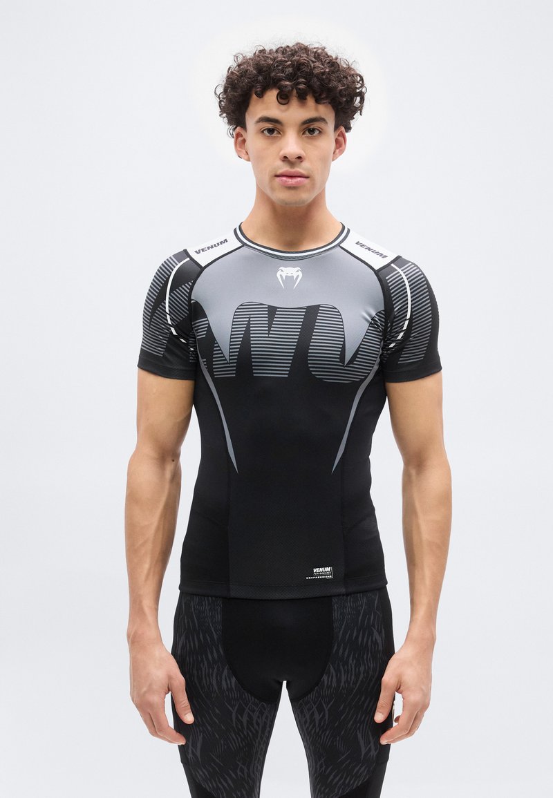 Νεαρός άνδρας με κατσαρά μαλλιά που φοράει στενό αθλητικό μπλουζάκι συμπίεσης σε μαύρο και γκρι χρώμα και κολάν με σχέδιο, στέκεται μπροστά σε λευκό φόντο.