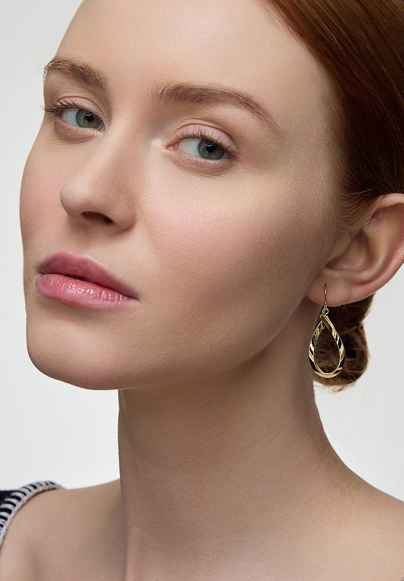 Złote kolczyki w kształcie kropli z zakręconym wzorem wiszą na haczyku, odbijając światło na gładkiej karnacji i neutralnym tle.