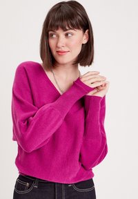 Pull-over en V magenta en matériau doux, doté de manches longues à bords côtelés et d'une coupe décontractée, associé à un jean foncé.