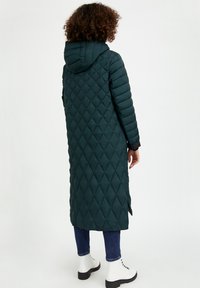 Abrigo largo acolchado de color verde azulado con capucha, con costuras en forma de diamante, aberturas laterales y puños negros, combinado con jeans azules y botas blancas.