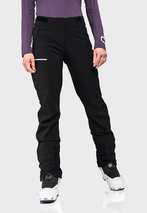 Persoon die zwarte skibroek draagt met ritszak en witte skischoenen, gecombineerd met een paarse longsleeve tegen een lichte achtergrond.