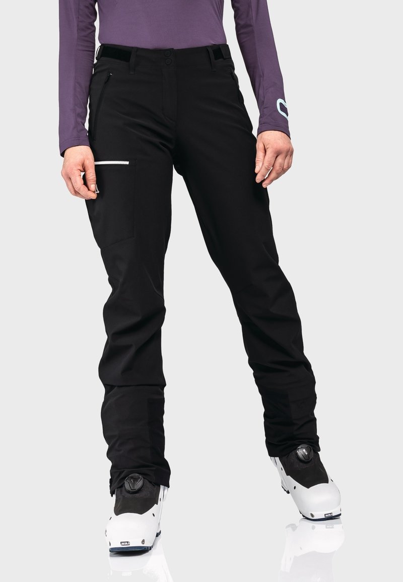 Personne portant un pantalon de ski noir avec poche zippée et des bottes de ski blanches, associé à un haut violet à manches longues sur fond clair.
