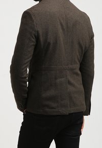 Brauner Wollblazer mit maßgeschneiderter Passform, seitlichem Rückenschlitz und Knöpfen an den Manschetten. Strukturierte Oberfläche, getragen über schwarzen Hosen.