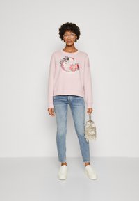 Sudadera rosa con bordado floral, corte holgado y cuello redondo. Combinada con jeans de mezclilla azul claro y zapatillas blancas, sosteniendo una bolsa plateada.
