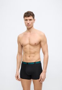 Μυώδης άνδρας με κοντά σγουρά μαλλιά, φορώντας μαύρα εσώρουχα Puma boxer, στέκεται μπροστά σε απλό λευκό φόντο, κοιτάζοντας μπροστά.