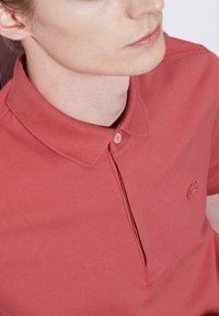 Röd pikétröja i texturerad tyg, ribbad krage och tvåknappsstängning. Har en diskret broderad logotyp på bröstet.