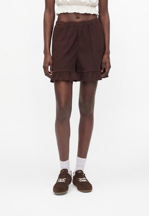 Femme portant un short marron foncé à volant dans l’ourlet, un top court blanc, des chaussettes blanches et des chaussures marron à lacets, debout devant un fond blanc.
