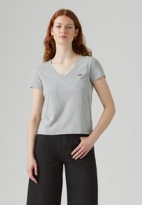 Gray cotton t-shirt with a v-neck and short sleeves. Features a small red logo on the left chest. Worn with black wide-leg pants.