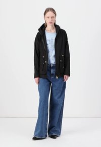 Μαύρο αδιάβροχο μπουφάν με κουκούλα, ρυθμιζόμενη μέση με κορδόνι και μπροστινές τσέπες, φορεμένο πάνω από ένα ανοιχτό μπλε μπλουζάκι και συνδυασμένο με φαρδιά μπλε τζιν.