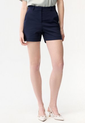 Kvinde iført marineblå, højtaljede, skræddersyede shorts og hvide slingback-hæle med spids tå, stående mod en ensfarvet hvid baggrund.