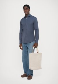 Chemise bleu marine à manches longues, jeans bleu clair et chaussures marron. Tenant un tote bag beige avec des anses marron. Tissu lisse et coupe décontractée.