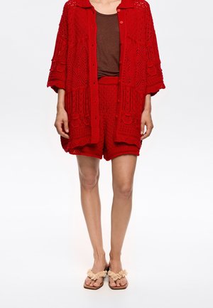 Red knitted open-front cardigan with textured patterns, paired with matching red shorts, worn over a brown top and beige sandals.