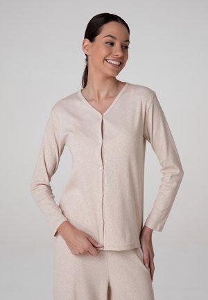Donna con capelli scuri legati indietro che indossa una maglia beige a maniche lunghe con bottoni e pantaloni abbinati, sorridente e che guarda di lato.