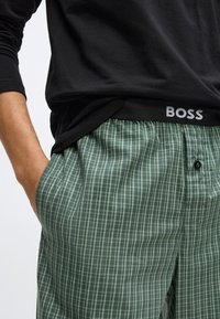 Cămașă neagră cu mâneci lungi asortată cu pantaloni verzi cu carouri. Pantalonii au o textură netedă, talie elastică și un detaliu cu nasture.