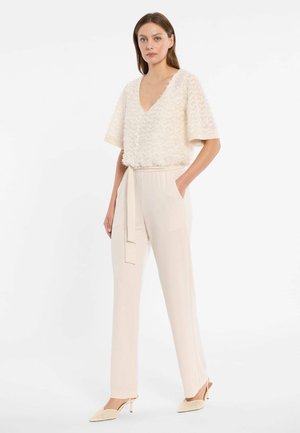 Femme debout portant un haut texturé à manches courtes crème avec col en V, un pantalon crème taille haute avec ceinture et des talons pointus crème.