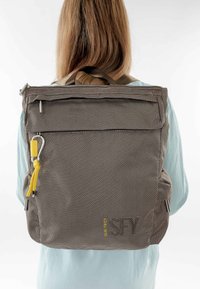 Žena v světle modré košili nesoucí čtvercový šedý látkový batoh s předním zipem a žlutým jezdcem.