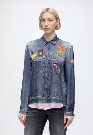 Jeune personne portant une veste en jean bleue avec des patches colorés et un jean noir, debout devant un fond clair uni.