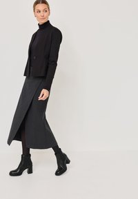 Černé padnoucí sako s jedním knoflíkem, spárované se šedou pruhovanou sukní s vysokým pasem a předním rozparkem, a černými koženými kotníkovými botami na podpatku.