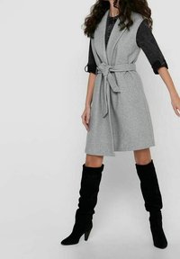 Vestito gilet in tessuto grigio con cintura annodata, caratterizzato da un colletto a revers e una lunghezza al ginocchio. Abbinato a stivali neri alti fino al ginocchio.