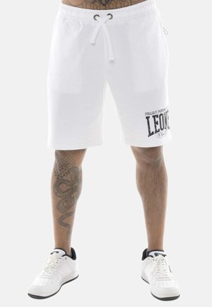 Man draagt witte shorts met trekkoord en het "Leone"-logo op het linkerbeen en witte sneakers, toont getatoeëerde benen.