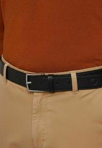 Czarny skórzany pasek z srebrną metalową klamrą, noszony na beżowych spodniach, zestawiony z rdzawej barwy bluzką. Pasek ma teksturowane wykończenie.