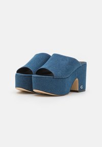 Mule in denim con zeppa, con punta aperta, di colore blu profondo, texture liscia e design arrotondato. Presenta un piccolo logo accentato sul tallone.