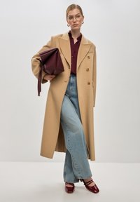 Vaaleanbeige villa-takkia, jossa on kaksinkertaisesti nepparit, maroonin värinen nappipaitapusero, vaaleansiniset farkut ja viinipunainen käsilaukku. Kiiltävät punaiset teräväkärkiset kengät.