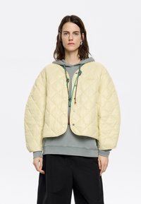 Gequilted, lichtgele cropped jack met een ronde vorm, voorzien van een drukknoopsluiting en contrasterende trekkoorden. Gecombineerd met een grijze hoodie eronder.