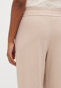 Béžové kalhoty s hladkým povrchem a elastickým pasem, které mají volný střih a jednoduchý design. Zobrazení ukazuje detaily zadní a boční strany.