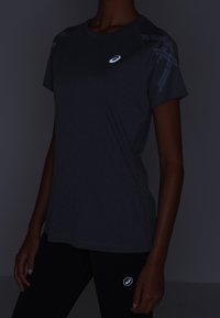 Camiseta deportiva gris con un logo reflectante y detalles de malla con patrón en los hombros, combinada con leggings negros.