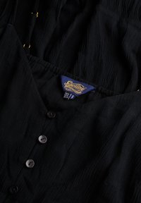 Zwarte textuurblouse met een knoopsluiting aan de voorkant, een elastische tailleband en een label met gouden details. De stof heeft een gekreukt uiterlijk.