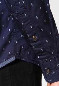 Navyblauwe shirt met kleine witte diamantpatronen, met knoopmanchetten en een gemêleerde stof. Heeft een contrasterende zoom met een blauwe binnenlaag.