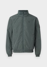 Kurze, mit Reißverschluss zu schließende Jacke in dunkelblaugrün, aus leichtem Stoff gefertigt. Sie verfügt über einen Stehkragen, elastische Bündchen und ein kleines Logo auf der Brust.