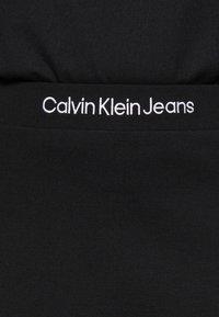 Černý pás Calvin Klein Jeans s bílým vyšitým názvem značky, hladká textura a elastický materiál. Detaily se zaměřují na jednoduchost a padnutí.
